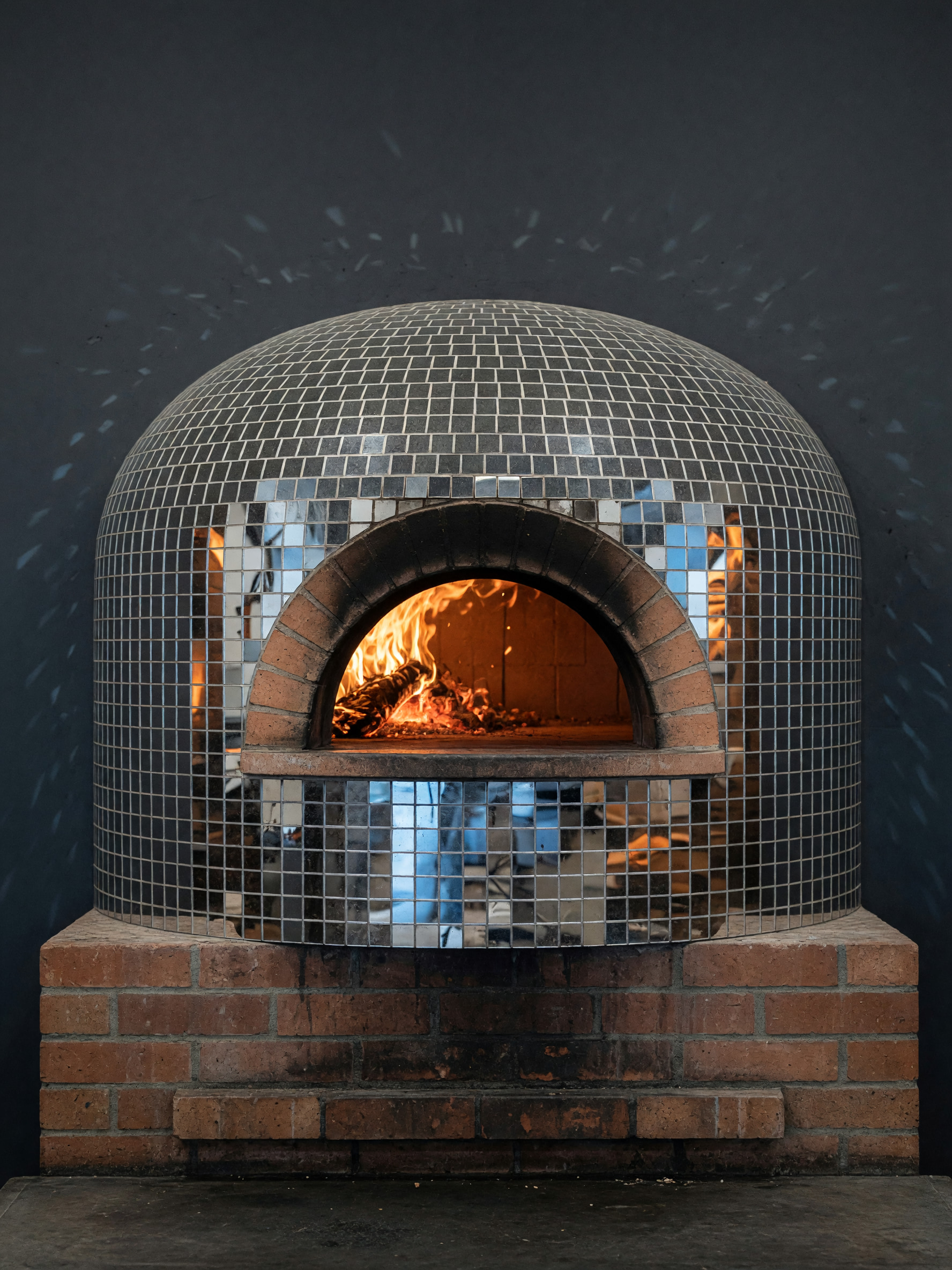 Holzbefeuerter Pizzaofen mit spiegelnder Discokugel-Kuppel und Feuerschein in der Ofenöffnung